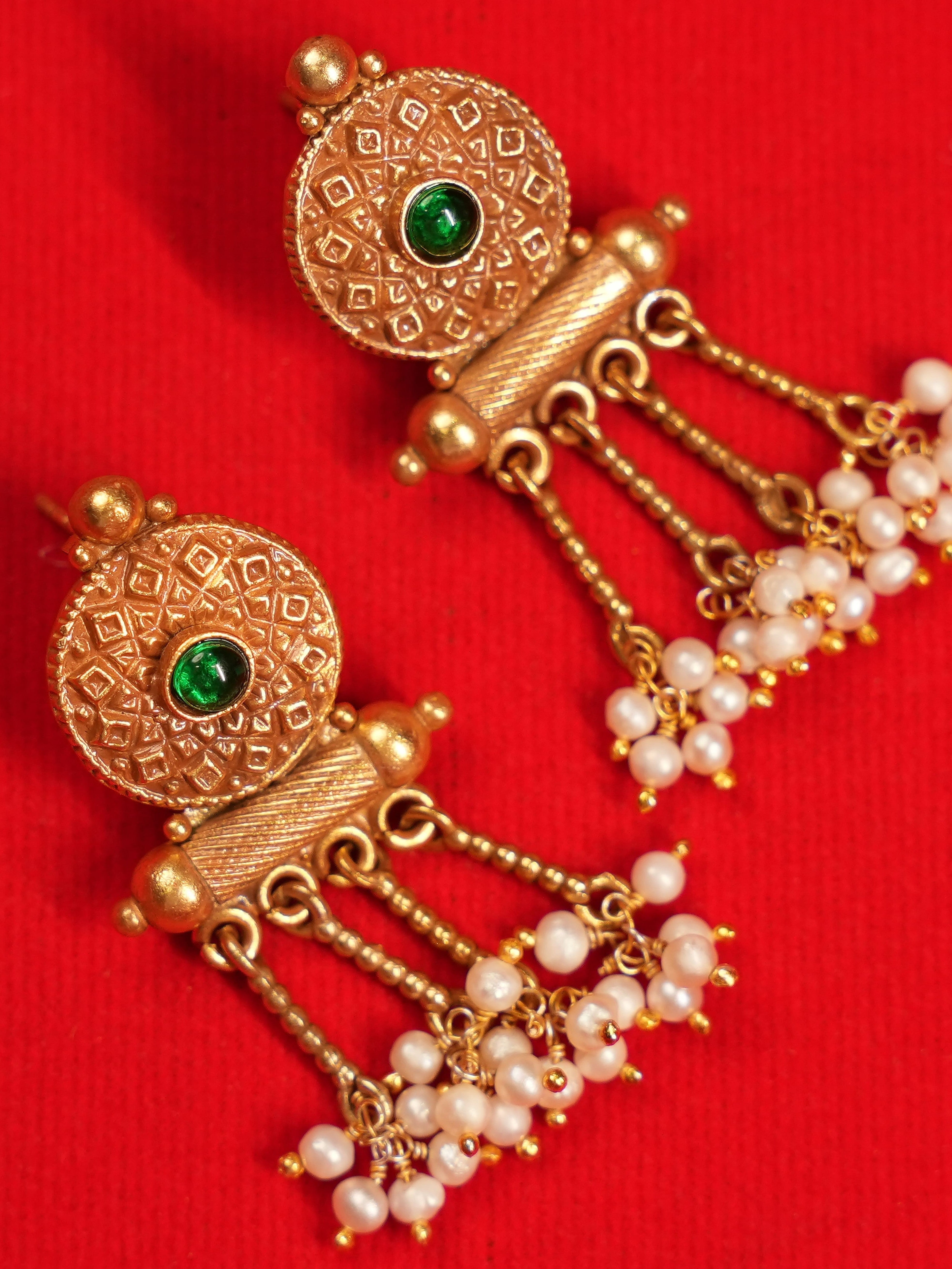 A pair of traditional Indian gold dangle earrings with green stone accents and white pearl beads, displayed on a textured red background.